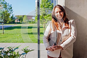 Happy confident office worker holding laptop by window. Caucasian young woman looking at camera. Business