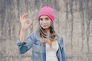 Happy confident smiling woman without make-up dressed in jeans shirt, white hat, with long curly hear showing ok-sign against grey