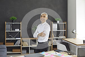 Happy confident successful senior businessman standing by office desk and smiling