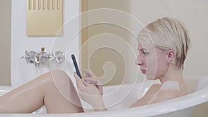 Happy confident woman typing on smartphone in bathtub. Side view portrait of blond beautiful Caucasian lady surfing