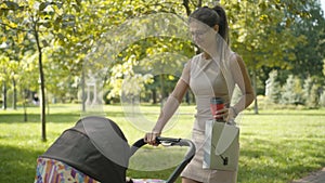 Happy confident young mother walking with baby stroller in sunny park. Portrait of Caucasian stylish businesswoman in