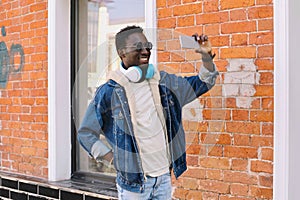 Happy cool smiling african man taking selfie picture by smartphone on city street over brick wall