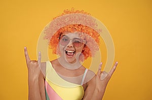 happy cool teen girl in sunglasses wearing orange curly wig hair on yellow background, childhood.