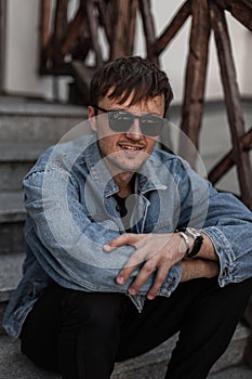 Happy cool young man hipster in stylish blue denim clothes in trendy sunglasses sits on a vintage staircase in the city