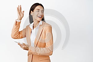 Happy corporate woman using smartphone, turn behind to wave at friend, saying hello to coworker while passing by with