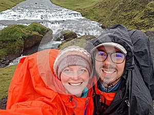 Happy couple adventure travelers man and woman in raincoats with waterfall behind. Freedom traveling and active lifestyle concept