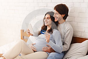 Happy couple awaiting baby and making online shopping on bed at home