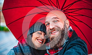 Happy couple of a bearded bald man and a caucasian brunette woman taking a selfie portrait holding an umbrella during christmas