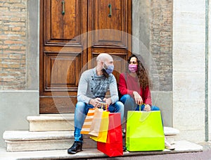 Happy couple of a bearded bolt man and a curly caucasian woman wearing face mask carrying shopping bags sitting on stairs in the
