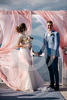 Happy couple, the bride and groom, stand on the pier under the wedding arch