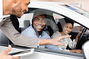 Happy Couple Buying Car Talking With Salesman Sitting In Auto