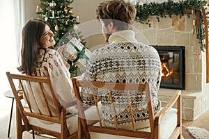 Happy couple in cozy sweaters exchanging stylish wrapped christmas gifts on background of fireplace with festive mantle and modern