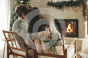 Happy couple in cozy sweaters kissing and exchanging stylish christmas gifts on background of fireplace with festive mantle and