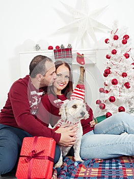 Happy couple with dog, all in Christmas clothes sitting near Christmas tree