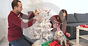 Happy couple with dog, all in Christmas clothes sitting near Christmas tree