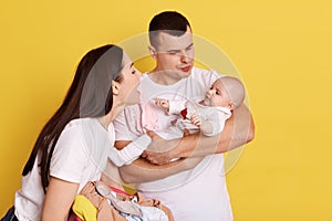 Happy couple embracing and looking at newborn child over yellow background, talking to little daughter with love and smile,