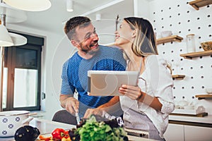 Happy couple is enjoying and preparing healthy meal in their kitchen and reading recipes on the digital tablet