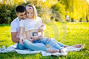 Happy couple expecting baby, pregnant young woman with husband sitting on grass and holding ultrasound pictures, young