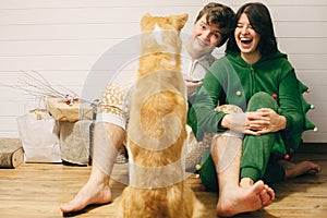 Happy couple in festive pajamas having fun and feeding their dog with canned food in room. Celebrating Christmas or New Year eve