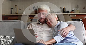 Happy couple grandparents cuddle on sofa look on laptop screen