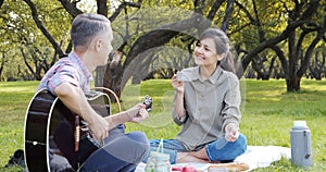 Happy couple with guitar having rest on picnic in the park on the lawn. Romance, vacation and summer concept.