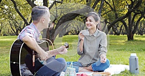 Happy couple with guitar having rest on picnic in the park on the lawn. Romance, vacation and summer concept