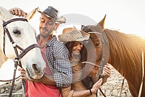 Happy couple having fun with horses inside stable - Young farmers sharing time with animals in corral ranch