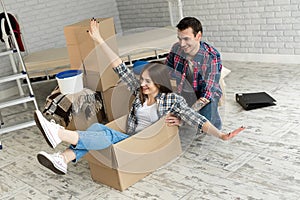 Happy couple having fun laughing moving into new home, excited woman riding sitting in cardboard box