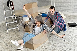 Happy couple having fun laughing moving into new home, young excited woman riding sitting in cardboard box while man