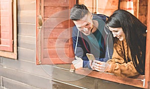 Happy couple having fun with smartphone inside camping wood house - Young people enjoying mountain vacation - Love, travel and