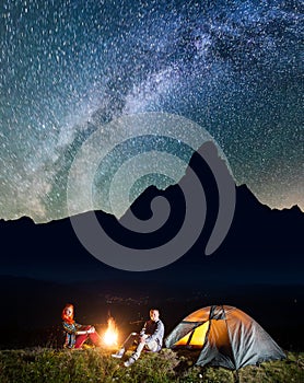 Happy couple hikers sitting near campfire and lighting tent under incredibly beautiful starry sky. Low light