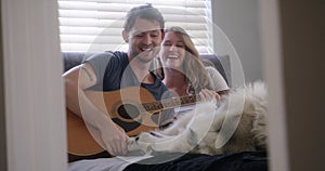 Happy couple, home and playing guitar with dog for love and bonding in bed with pet. People with animal, playful husky