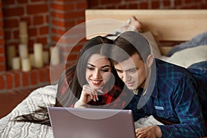 Happy couple with laptop spending time together at home, browsing internet in bed, smiling and having fun