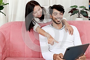Happy couple in living room, Asian female hugging around her boyfriend neck while he using tablet on sofa. Lover showing love