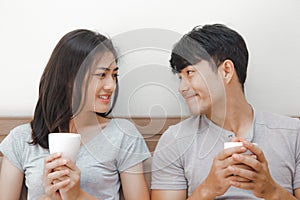 Happy couple in love. asian young man and woman holding coffee cup on the bed. The lover smile and look at each other face happily