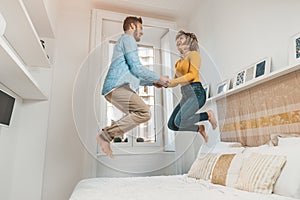 Happy couple in love having fun jumping on bed at home. Handsome man and beautiful woman are enjoying