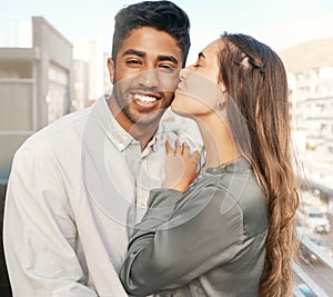 Happy couple, love and kiss on cheek from girlfriend bonding with boyfriend standing on a city building balcony on a