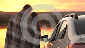 Happy couple in love, travelers man, woman cover themselves with blanket next to car and admire beautiful sunset on