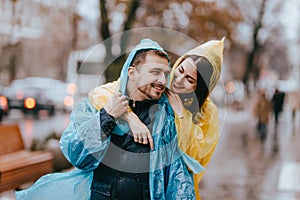 Happy couple loving guy and his girlfriend dressed in raincoats are hugging on the street in the rain