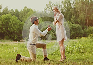 Happy couple, man kneeling down and proposing ring to his woman outdoors on the grass, wedding concept