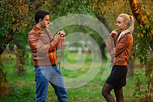 Happy couple, man and woman shooting on the old camera outdoor in the green autumn park in the red bomber leather coats