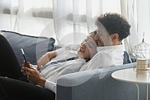 happy couple middle aged man and woman hugging lying digital tablet relaxing on sofa in modern house living room. browsing