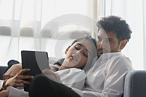 happy couple middle aged man and woman hugging lying digital tablet relaxing on sofa in modern house living room. browsing