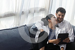 happy couple middle aged man and woman hugging lying digital tablet relaxing on sofa in modern house living room. browsing