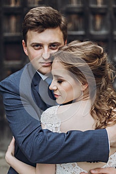 Happy couple of newlyweds posing near ancient wooden castle gates, handsome smiling groom embracing beautiful blonde bide in lace