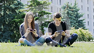 Happy couple play mobile game on smartphone, sitting on the grass in the park. Man win the game, woman lost.