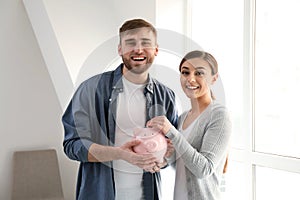 Happy couple putting coin into piggy bank indoors. Money savings concept