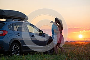 Happy couple relaxing beside their SUV car during honeymoon road trip at sunset. Young man and woman enjoying time