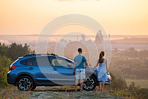 Happy couple relaxing beside their SUV car during honeymoon road trip at sunset. Young man and woman enjoying time together