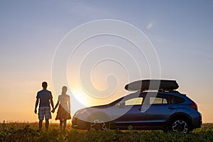 Happy couple relaxing beside their SUV car during honeymoon road trip at sunset. Young man and woman enjoying time together